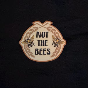 May include: A sticker with a cream-colored background and a brown border. The sticker features two bees and the text "NOT THE BEES" in black. The bees are yellow and black, and the design includes brown wheat stalks.