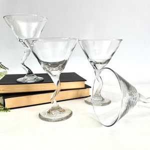 May include: Three clear glass martini glasses with twisted stems. The glasses are arranged on a stack of books with a green plant in the background.