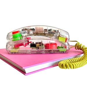 May include: A clear, retro-style telephone with visible internal components rests on a pink book. The phone features a yellow coiled cord and colorful accents, including red and green. The design evokes a vintage aesthetic.