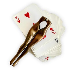 May include: A bronze-colored nutcracker in the shape of crossed legs rests on a deck of playing cards. The cards are white with red heart symbols and black numbers and letters. The nutcracker has a smooth, polished finish.