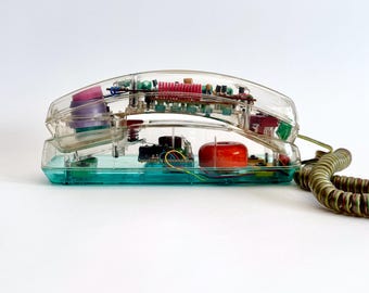 May include: A clear, transparent telephone with visible internal components. The base is a teal color, and the handset features red, pink, and green elements. The coiled cord is a muted olive green.