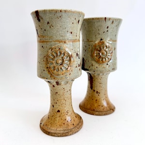 May include: Two handmade ceramic goblets with a speckled, earthy glaze. Each goblet features a pedestal base, a cylindrical midsection with a floral medallion, and a flared rim. The color palette includes shades of tan, brown, and gray.