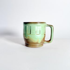 May include: A ceramic mug featuring a light mint green glaze on the upper portion and a solid brown base. The mug has a vertical ribbed texture around the top half and a sturdy, angular handle extending from the right side of the vessel.