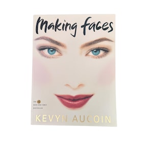 May include: A book cover with the title "Making Faces" in black script above a woman's face. The woman has blue eyes, rosy cheeks, and red lipstick. The author's name, Kevyn Aucoin, is printed in gold below.