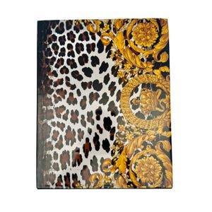 May include: A rectangular notebook with a glossy cover featuring a split design. The left side displays a black and brown leopard print pattern on a white background, while the right side showcases an ornate gold floral and crown motif.