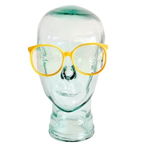 May include: Yellow-framed eyeglasses with a round shape are displayed on a clear glass head. The glasses have a bright yellow color and a classic, oversized design. The clear lenses are visible through the frames.