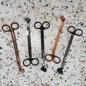 May include: Five candle wick trimmers, two are rose gold, two are black, and one is silver. The trimmers are made of metal and have a long handle with a small scissor-like head.