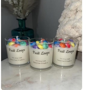 May include: Three clear glass candles with white wax and colorful, fruit-shaped wax pieces on top. The candles have the text "Fruit Loops" printed on them. The candles are on a white surface.