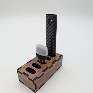 May include: A wooden organizer with three slots, holding a black vape pen and a vape pod. The organizer is brown with black edges. The vape pen has a diamond pattern and a heart-shaped cutout.