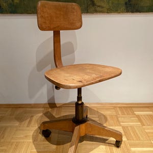 Könnte beinhalten: Vintage Bürostuhl aus Holz mit geschwungener Rückenlehne und Sitzfläche. Der Stuhl hat ein Fünf-Sterne-Gestell mit Rollen. Das Holz hat eine natürliche Oberfläche. Die Höhe des Stuhls ist verstellbar. Höhe ca. 70 cm.