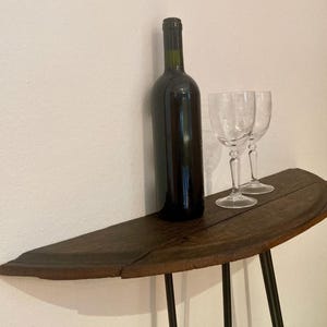 Könnte beinhalten: Eine dunkle Weinflasche und zwei klare Weingläser stehen auf einem rustikalen, halbkreisförmigen Holztisch mit schwarzen Metallbeinen. Der Tisch steht vor einer weißen Wand und schafft eine schlichte, elegante Präsentation.