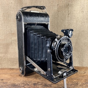 Könnte beinhalten: Eine schwarze Vintage-Kodak-Kamera mit Ledergriff und faltbarem Balgen. Die Kamera hat ein silbernes Objektiv und einen kleinen Blitzaufsatz. Auf dem Objektiv steht "Kodak Anastigmat" und "f=10,5cm". Die Kamera ist geöffnet und einsatzbereit.