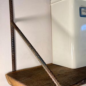 Könnte beinhalten: Ein rustikales Holzregal mit einer Metallhalterung. Die Halterung besteht aus Betonstahl und hat eine rostige Oberfläche. Das Regal ist an einer weißen Wand montiert.