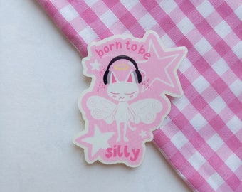 Könnte beinhalten: Ein rosa Aufkleber mit einer weißen Katze mit Engelsflügeln, Heiligenschein und Kopfhörern. Der Aufkleber trägt die Aufschrift "born to be silly" und Noten. Der Hintergrund ist ein rosa-weißes Vichy-Muster.