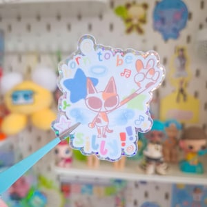 Könnte beinhalten: Ein holografischer Aufkleber mit einer Cartoon-Katze mit orangefarbenen und weißen Markierungen, Flügeln und dem Schriftzug „born to be silly“. Der Aufkleber enthält einen blauen Stern, Noten und ein Friedenszeichen.