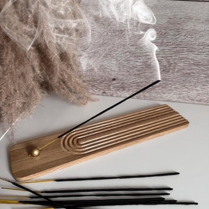 Può includere: Un porta incenso in legno con un accento dorato e un design scanalato. Un bastoncino di incenso nero sta bruciando e rilasciando fumo. Un mazzo di bastoncini di incenso neri si trova su una superficie bianca.