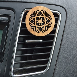 Può includere: Un profumatore per auto in legno con un design geometrico. Il profumatore è marrone e ha un centro nero. È appeso alla bocchetta di ventilazione di un'auto.