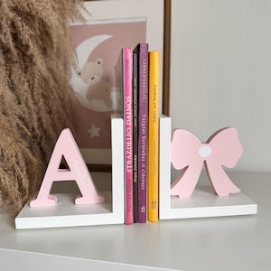 May include: A pair of pink and white wooden bookends. One is an "A" with "Aurelia" on it, the other a bow. Books of different colours are placed between them. A framed picture with a moon and bear is in the background. The bookends are for books.