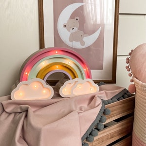 Könnte beinhalten: Ein hölzernes Regenbogen-Nachtlicht in den Farben Rosa, Grün, Gelb und Weiß. Der Regenbogen hat auf jeder Seite eine leuchtende Wolke. Der Regenbogen steht auf einer rosa Decke mit grauem Besatz.