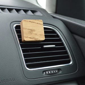 Può includere: Un deodorante per auto in legno con finitura in legno naturale. Il deodorante è posizionato in una bocchetta di ventilazione dell'auto.