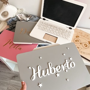 以下が含まれることがあります： キーボード付きのグレーのラップトップ、Milaという名前のピンクのノート、Hubertoという名前と星のアクセントが付いたグレーのノートなど、木製のおもちゃのコレクション。クマのイラストが描かれた木の板も表示されています。