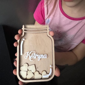 May include: Wooden mason jar-shaped plaque with the name "Kotryna" in white script. The jar contains wooden heart shapes. The plaque is held by a person wearing a pink shirt.