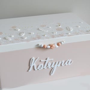 May include: A white wooden keepsake box with a pink lid. The lid is decorated with a floral design and a string of wooden beads. The name "Kotryna" is written in white letters on the front of the box.