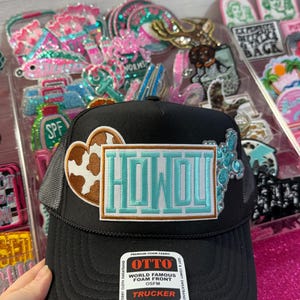 Puede incluir: Gorra de camionero negra con la parte trasera de malla y un parche que dice "HOWDY" en letras turquesas. El parche también presenta un corazón de piel de vaca y una cruz decorativa. La gorra tiene un cierre a presión y una etiqueta "TRUCKER".