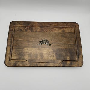 May include: A rectangular wooden tray with a dark brown finish. The tray features a recessed border and a decorative metal lotus flower emblem. A small, rectangular cutout is visible on the bottom edge.