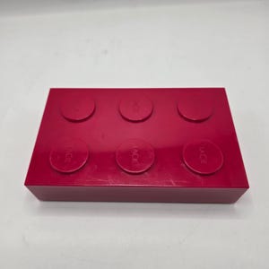 May include: A bright red rectangular storage container with six circular raised elements on the top. The word "LACIE" is printed on each of the circular elements. The container is made of a solid, smooth material.