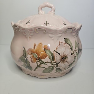 May include: A pink ceramic lidded jar with a floral design. The jar features a handle on the lid and is decorated with orange and white flowers and green leaves. Gold accents are present on the rim.