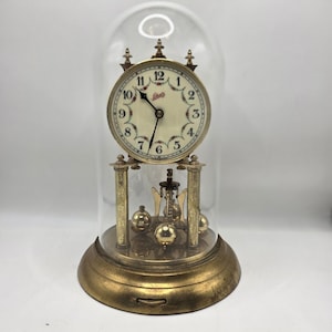 May include: An antique clock with a cream-colored face, black hands, and Roman numerals, encased in a glass dome. The clock has a gold-colored base and decorative elements, including a pendulum with three gold-colored spheres.