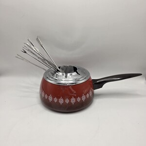 May include: A red fondue pot with a black handle and a silver lid. The pot has a decorative white pattern around the base and includes several fondue forks. This kitchenware is ideal for serving melted cheese or chocolate.