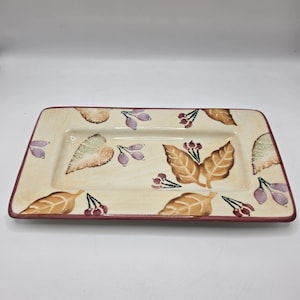 May include: Rectangular ceramic serving platter with a cream-colored background and a burgundy rim. The platter is decorated with brown and tan leaves, and clusters of purple berries.