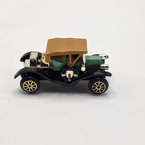 May include: A miniature vintage-style automobile featuring a metallic green body, a tan roof, and black fenders. The vehicle includes gold-colored wheels with spokes, a gold-toned front radiator, and a spare tire mounted on the side.