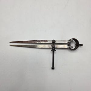 May include: A vintage metal caliper tool with pointed tips and a screw adjustment. The caliper has a rusty appearance, suggesting age and use. The tool is designed for measuring distances and diameters, commonly used in engineering and crafts.