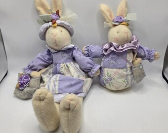 May include: Two plush rabbit dolls with cream-colored fabric bodies. The larger rabbit wears a lavender dress with floral and striped patterns, a matching bonnet, and carries a small mesh bag. The smaller rabbit wears a lavender ruffled collar.