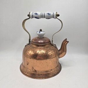 May include: A copper tea kettle with a white and blue floral patterned handle and lid. The kettle has a spout and a curved handle. The copper has a speckled appearance. The kettle is designed for stovetop use.