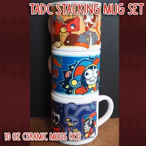 Könnte beinhalten: Ein Set aus drei stapelbaren Keramikbechern mit bunten Cartoon-Designs. Jeder Becher fasst 296 ml. Die Becher zeigen lebendige Charaktere und Farben. Der Text "TADC STACKING MUG SET" und "10 OZ CERAMIC MUGS (X3)" ist auf dem Bild zu sehen.