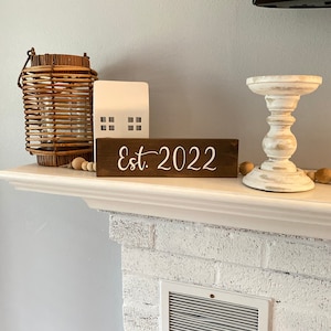 Könnte beinhalten: Ein Holzschild mit dem Text "Est. 2022" in weißen Buchstaben auf braunem Hintergrund. Das Schild steht auf einem weißen Kaminsims mit einer Weidenlaterne, einem weißen Keramikhaus und einem weißen Kerzenhalter.