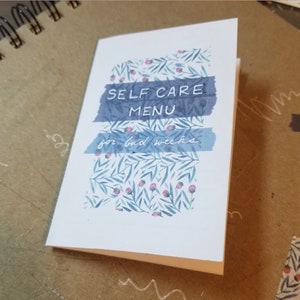 May include: A white card with a blue and pink floral pattern. The card has the text "SELF CARE MENU for bad weeks" written on it.