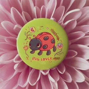 May include: A round lime green pin-back button featuring a red ladybug with black spots, surrounded by small floral illustrations and the text "BUG LOVER" printed in red. The design includes delicate line art of flowers and leaves.