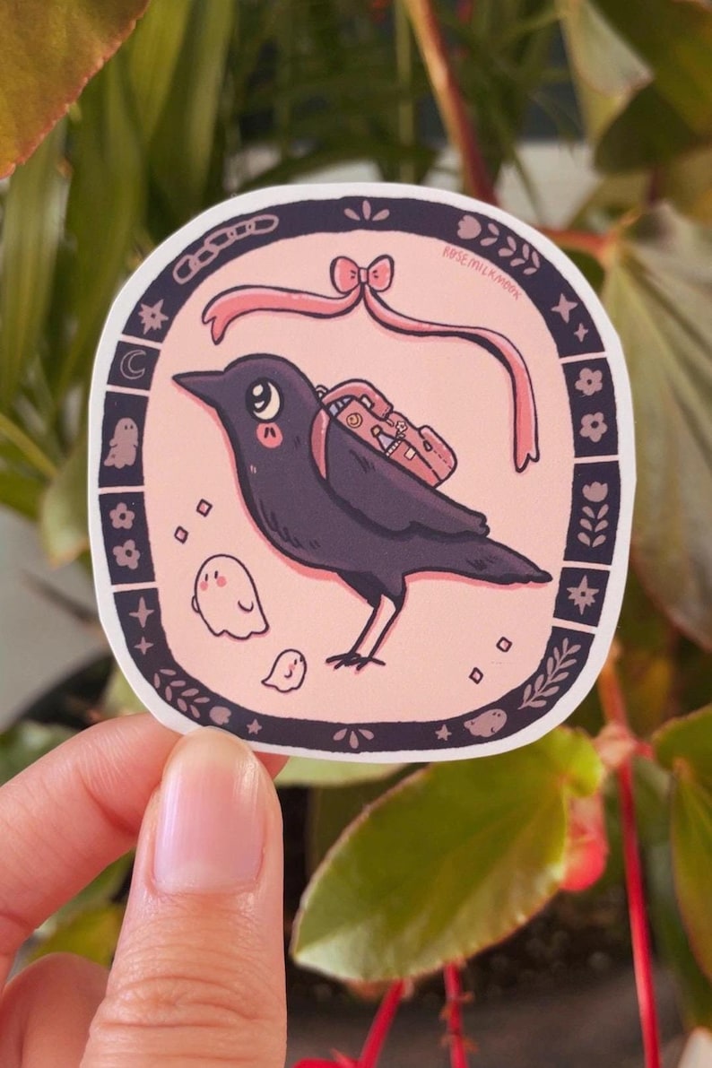 May include: A pink and black sticker with a cartoon crow wearing a backpack and a pink ribbon. The crow is surrounded by small white ghosts and stars. The sticker says "Rose Milkmoon" in the top right corner.