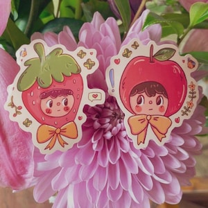May include: Two stickers featuring cartoon characters dressed as a strawberry and an apple. The strawberry character is red with a green stem, and the apple character is red with a green leaf. Both have orange bows and are surrounded by decorative elements.