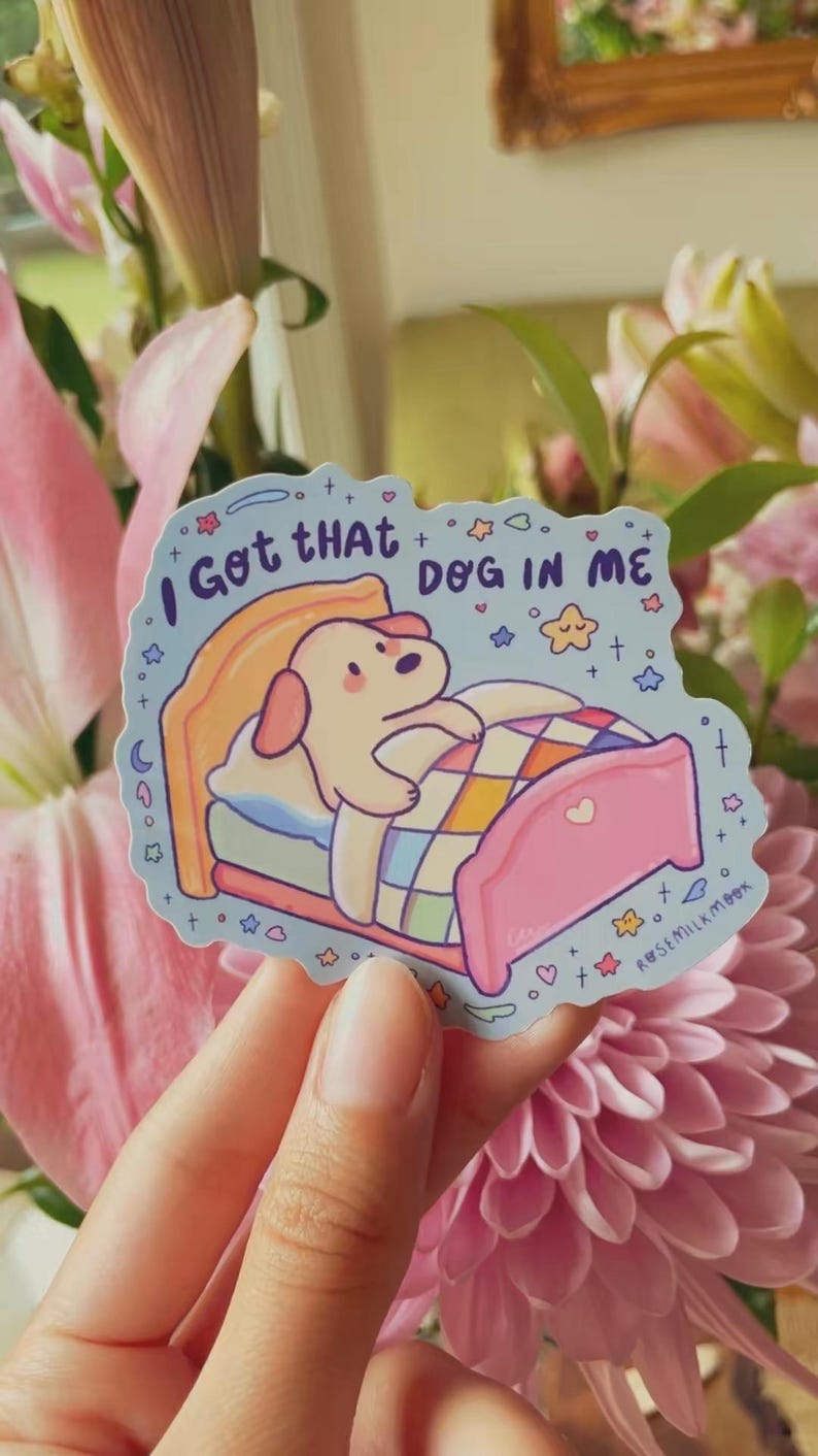 May include: A die-cut sticker featuring a light-colored cartoon dog tucked into a pink bed with a colorful checkered blanket. The light blue background includes stars, hearts, and the text "I Got tHAt DOG IN ME" along with "ROSEMILKMOOK".