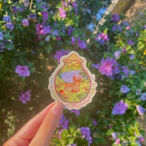 May include: A whimsical sticker featuring a deer resting in a meadow, enclosed within a decorative, scalloped border. The sticker has a soft color palette with shades of pink, green, and purple. The background shows a lush garden with purple flowers.