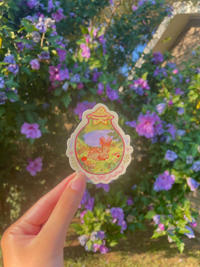 May include: A whimsical sticker featuring a deer resting in a meadow, enclosed within a decorative, scalloped border. The sticker has a soft color palette with shades of pink, green, and purple. The background shows a lush garden with purple flowers.