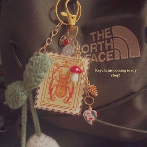 May include: A collection of keychains with various charms, including a crochet flower, a stamp-like image of a beetle, a mushroom, and a heart-shaped charm. The keychains are attached to a gold-colored clasp. Text on the image reads "Keychains coming to my shop!"