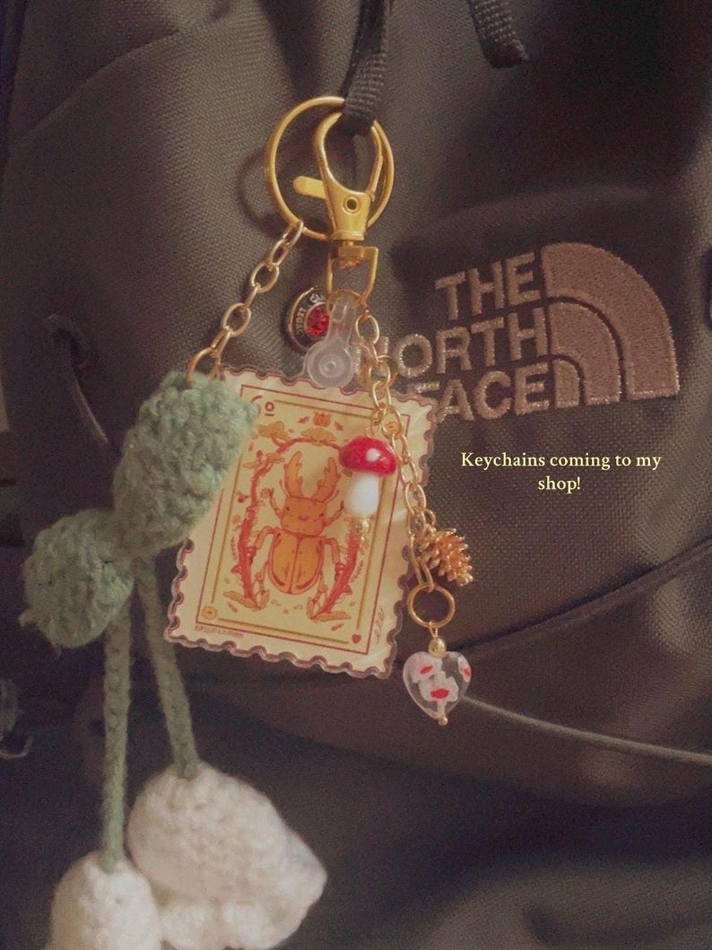 May include: A collection of keychains with various charms, including a crochet flower, a stamp-like image of a beetle, a mushroom, and a heart-shaped charm. The keychains are attached to a gold-colored clasp. Text on the image reads "Keychains coming to my shop!"