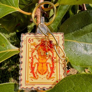 May include: A rectangular keychain featuring a stamp design with an orange beetle illustration, the number 6, and the text "ROSEMILKMOON". The keychain has a gold-colored ring, chain, and charms, including a red leaf and a ladybug.
